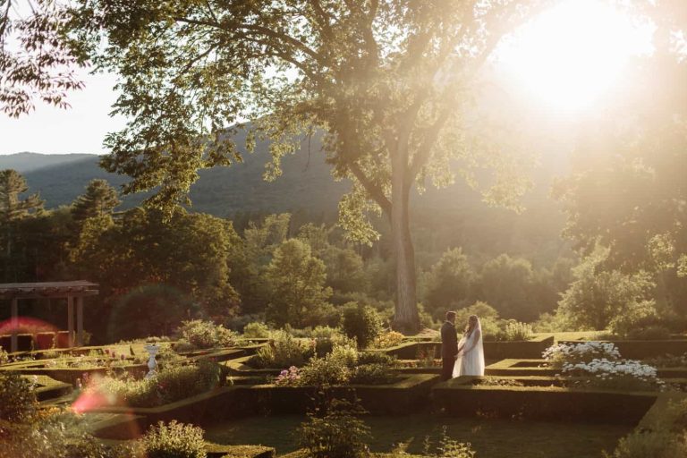 Hildene Wedding: bride and groom standing in a sunlit formal garden beneath a large tree with mountains in background.