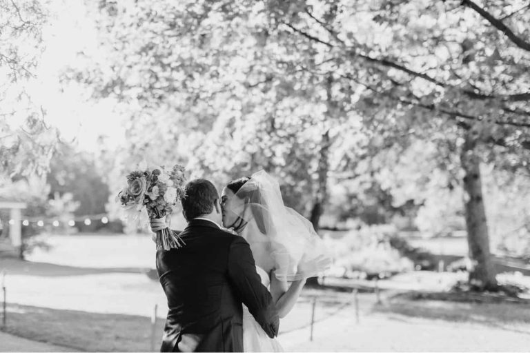 Bride and groom kissing under trees at a Basin Harbor wedding, bride's veil flowing and bouquet raised.