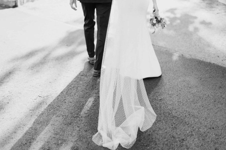 Bride and groom walking away on a sunlit pavement, bride's dotted veil trailing and bouquet in hand — Woodstock Inn wedding.