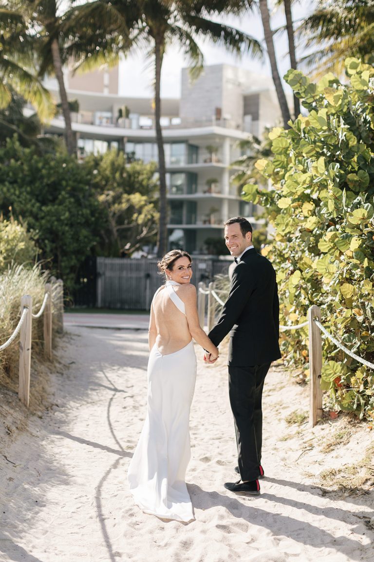 Bride in backless white gown and groom in black tux holding hands walking on a sandy path at a Miami Beach EDITION wedding.